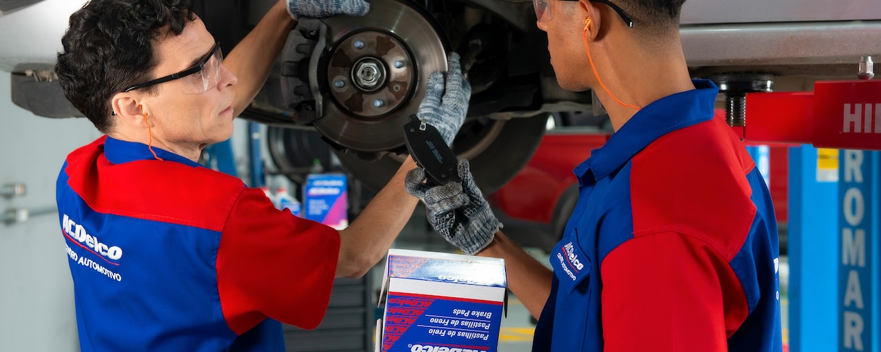 Dos mecánicos revisando frenos y pastillas ACDelco en vehículo elevado.